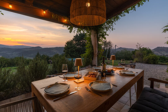 View of a dining table set for dinner on a terrace overlooking the rolling hills at sunset, with warm lighting and wine glasses, Smerillo, Marche, Italy.