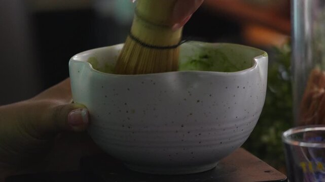 4K Close-up of hand whisking green matcha tea powder in ceramic bowl with traditional bamboo whisk at cafe, Japanese tea ceremony, matcha preparation, healthy drink and authentic culture.