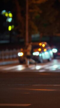 A pedestrian walks along a city street at night, surrounded by moving vehicles under streetlights, with urban buildings lining the road, creating a constant flow of nighttime city traffic