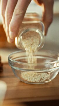 A man's hand tilts a glass jar, pouring dried white sesame seeds into a clear bowl on a tabletop, features precise ingredient handling for food preparation, linked to agricultural seed use