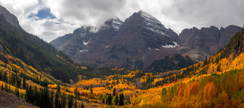 View of the Maroon Bells peaks rising above a vibrant valley filled with golden autumn aspen trees and evergreen forests in Pitkin County, Colorado, United States.
