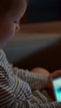 A child snuggles on a sofa, eyes fixed on a smartphone screen displaying bright cartoons, in a dim evening room, showing typical childhood screen use and the habit of late-night gadget viewing