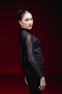 Full-length editorial portrait of a young Asian woman in a sheer black top and pants, touching her hair with a striking gaze against a deep red background.
