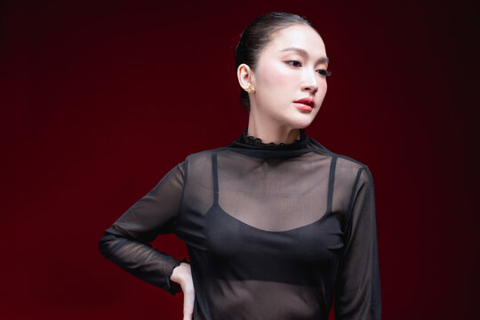 Confident beauty portrait of a young Asian woman in a sheer black top with hands on hips, gazing directly at the camera against a deep red background.