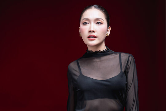 Confident beauty portrait of a young Asian woman in a sheer black top with hands on hips, gazing directly at the camera against a deep red background.