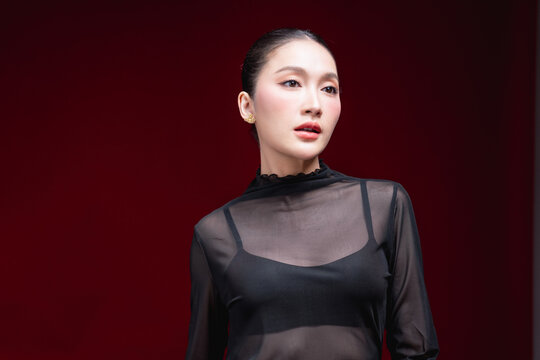 Side-profile beauty portrait of a young Asian woman in a sheer black top looking off to the side with a subtle expression against a deep red background.