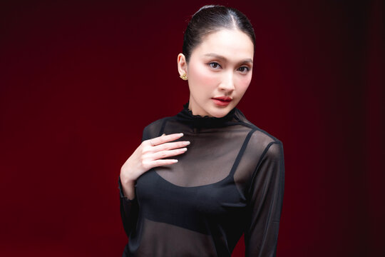 Side-profile beauty portrait of a young Asian woman in a sheer black top looking off to the side with a subtle expression against a deep red background.
