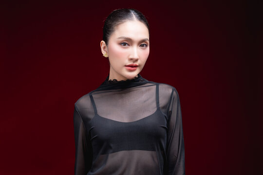 Cheerful close-up beauty portrait of a young Asian woman in a sheer black top laughing warmly with a raised hand near her face against a deep red background.