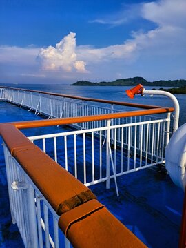 Ocean view from a ship deck with vibrant orange railings, calm blue sea, lush tropical islands, and dramatic white clouds under a bright blue sky.
