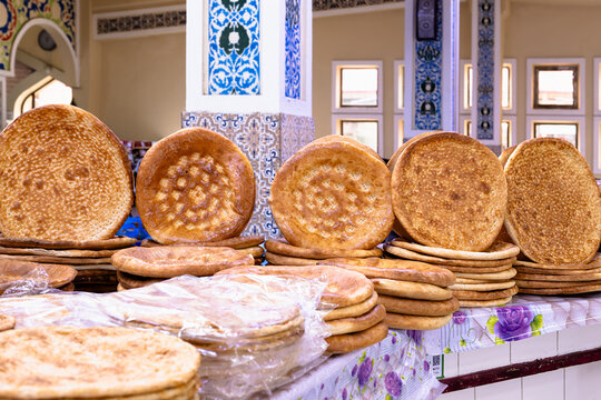Traditional Uzbek flatbread stacked in abundance at a bazaar counter in Bukhara. Golden round loaves with decorative stamped patterns displayed against ornate tilework in a market hall.