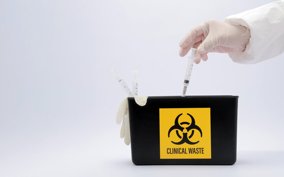 Cautious medical worker hand disposing syringe into safe clinical waste container with serious focused biohazard symbol indoors