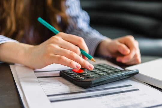 Female accountant using calculator and pencil while reviewing financial documents on a desk with printed reports and a notepad in a modern office setting