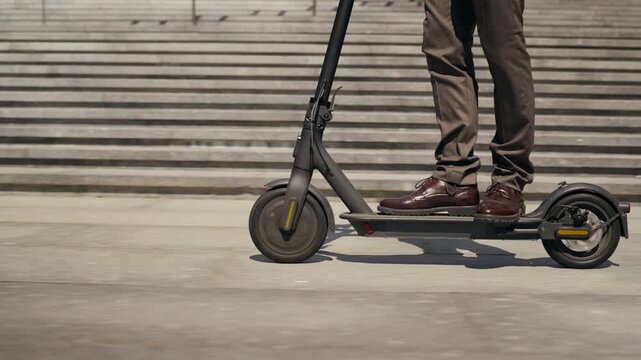 Unrecognizable businessman riding electric scooter on city street, tracking shot in slow motion. Perfect for commercial videos promoting eco-friendly urban commuting and modern business lifestyle