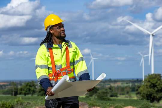 African american and caucasian engineers with blueprint and laptop checking wind turbines at windmill field farm. workers working at wind turbines farm. Alternative eco energy, environmental friendly