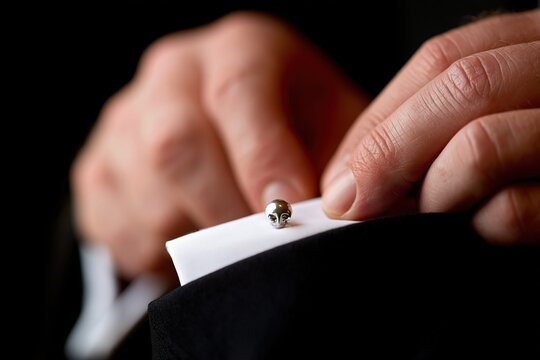 Close-up of a person's hand adjusting a silver skull cufflink on a white shirt cuff against a black suit jacket background
