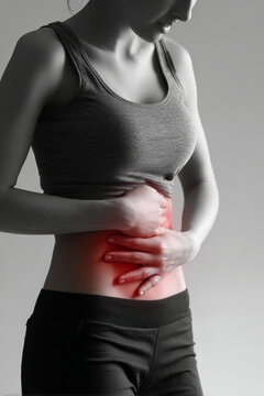 A woman stands in a simple space while holding her stomach with both hands. She appears to feel discomfort and is wearing a tank top and pants
