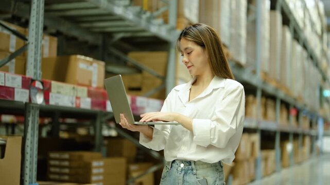 Young Asian female warehouse manager using laptop to check inventory among large shelves of cardboard boxes in a distribution center. Logistics, supply chain, and e-commerce business concept.