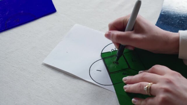 A woman wearing a wedding band traces the number 3 on a piece of green stained glass laid over a heart template during an art class.