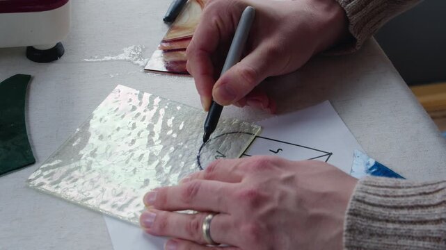 A man wearing a wedding band uses a sharpie to trace a piece of clear stained glass on a heart template during an art class.