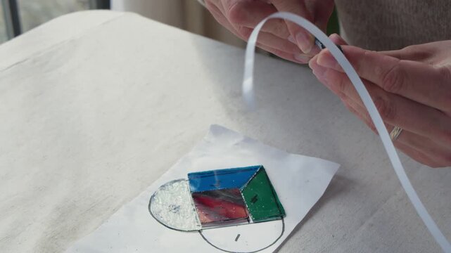 A man applies freshly peeled copper tape to a piece pf cut blue stained glass during an art class. He is sitting by a window and his full project is visible in the frame.
