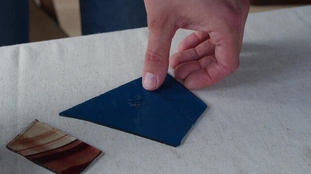 A man uses a scoring tool to follow a line drawn on a small piece of blue glass. He then places the tool down on the table and begins to pick up the piece.