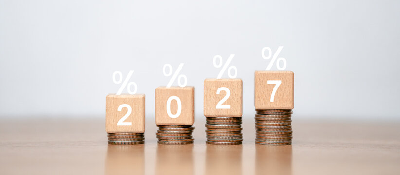 The wooden block with the number 2027 on top of a continuously rising stack of coins signifies planned growth in interest rates or accumulated financial returns.