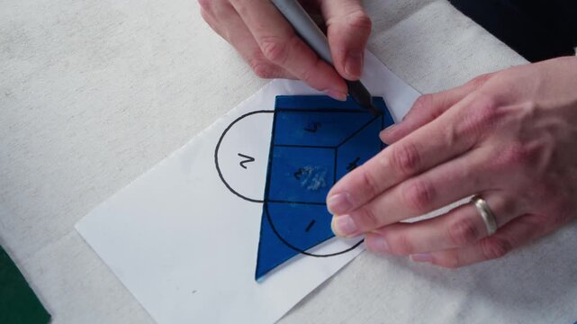 A man wearing a wedding band traces a piece of blue stained glass over one section of a heart template. He is in art class and using a sharpie to outline where he will cut the glass.