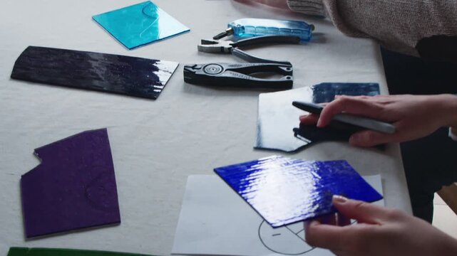 Two students work on a couples art project during a stained glass art class. The woman grabs a piece of blue glass and the camera follows her in for a close up of a heart template.