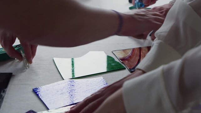 A teacher and her students are working on stained glass projects at a table in the sun. The table is full of colored glass and everyone reaches for tools or materials.
