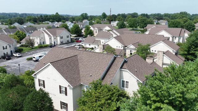 Beige townhouses with brown shingled roofs cluster around parking lot in Elizabethtown, Pennsylvania, dense tree canopy borders quiet residential streets nearby. Aerial drone orbit shot.