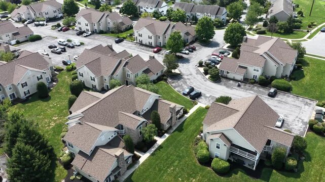 Cluster of townhome rooftops with tan asphalt shingles and steep gables surrounds paved parking court, driveways and green lawns in small American town, Pennsylvania. Aerial drone flyover shot