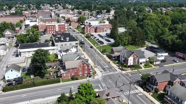Crosswalk-lined intersection cuts through historic downtown district in Elizabethtown, Pennsylvania, brick buildings and church spire border tree-shaded main street. Aerial drone approaching shot