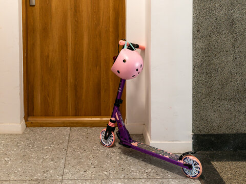 Purple children's kick scooter with a pink protective helmet parked beside a wooden apartment door. Practical urban family scene illustrating child mobility, outdoor recreation equipment, safety gear,