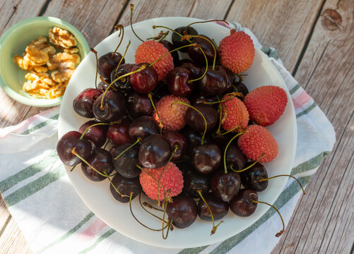 Dark sweet cherries, Prunus avium, and pink lychee fruit, Litchi chinensis, fill a white ceramic plate beside walnut kernels. Contrasting colors and natural sunlight create a vibrant food image for ex