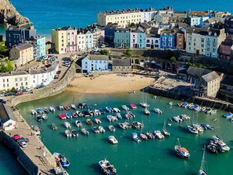 High-angle view of Tenby waterfront, harbor walls enclosing small beach and flotilla of dinghies, yachts, and motorboats under summer sunlight, Wales, UK