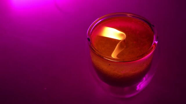 A close-up high-angle shot of a burning flame inside a clear glass candle holder. The warm yellow fire reflects in the liquid wax, illuminated by vibrant purple studio lighting on a flat surface.