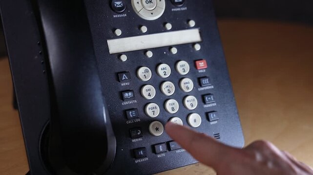 A close-up shot of a human hand using an index finger to dial a number on a black office landline telephone. The telecommunications device rests on a wooden desk inside a dimly lit workspace.