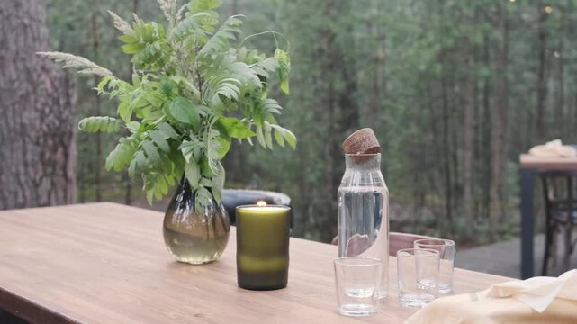 wooden table set with lit candle and fresh green branches in vase at forest cafe. water carafe with cork and glasses decorate outdoor dining table under string lights in pine woodland.