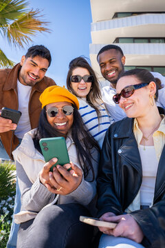 Vertical. Multicultural friends sit together and look at smartphones outdoors in a modern urban setting, sharing content and enjoying a connected social moment.