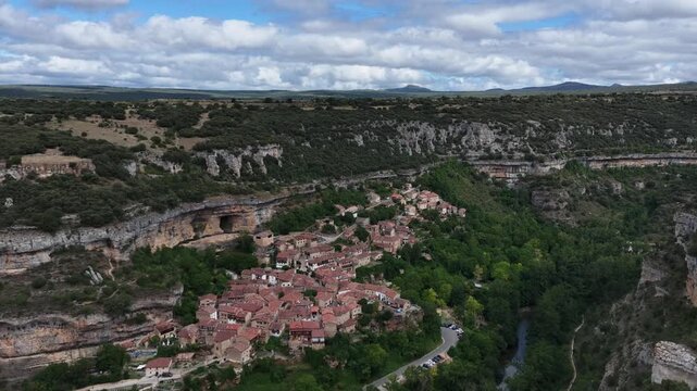 peaceful aerial view of a small village nestled within forested hills in rural, mountainous terrain with red-tiled roofs and stone buildings, Risco los camellos, Cascada de Orbaneja del Castillo