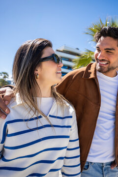 Vertical. Smiling man and woman share an affectionate moment outdoors, walking together in a modern neighborhood with palm trees and contemporary buildings.