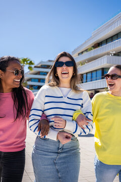 Vertical. Three women walk together arm in arm through a sunny modern neighborhood, enjoying friendship, conversation and an easygoing urban lifestyle.