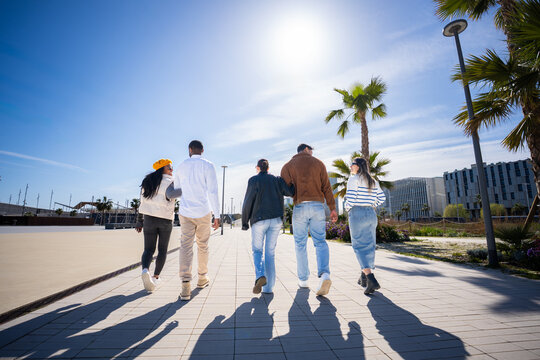 Group of friends walk away along a wide pedestrian promenade in a blue sky bright urban residential district with palm trees and clear blue sky.