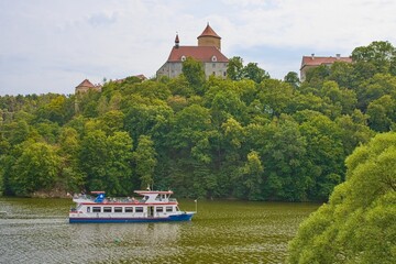 Scenic view of a passenger tour boat navigating the water of Brno Reservoir (Prygl) with the historic Veveri Castle rising above dense green forest on a hill. Concept of summer travel, water transport © Igor