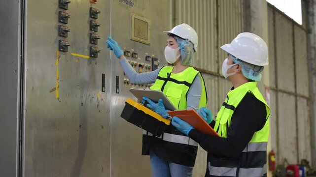 Professional engineers in safety helmets and face masks inspect an electrical control panel with clipboard checklist