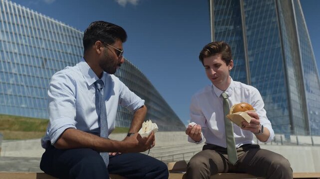 Caucasian and South Asian young adult businessmen sitting outdoors, eating lunch and discussing work during break. Low angle shot ideal for corporate culture and work-life balance concepts