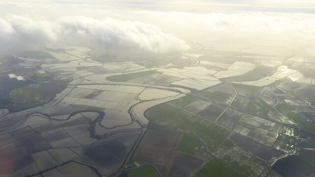 View from an airplane window shows flooded farmland and winding river patterns beneath soft clouds and diffused light.
