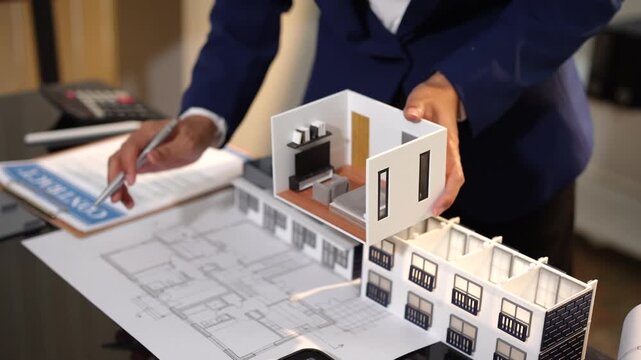Close-up of an architect or agent showing a modern house model over floor plans in an office, suggesting home design, real estate planning, and property presentation.