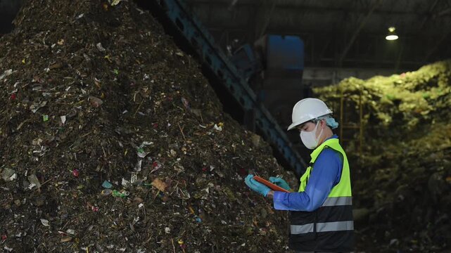 Environmental engineer in protective safety gear inspecting processed waste and taking notes on a clipboard at industrial recycling facility.