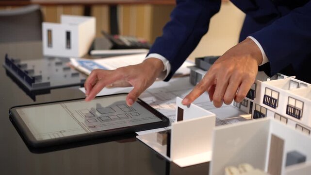 Architect reviewing digital floor plans on a tablet beside small building models in an office, showing design work, planning, and modern construction ideas.